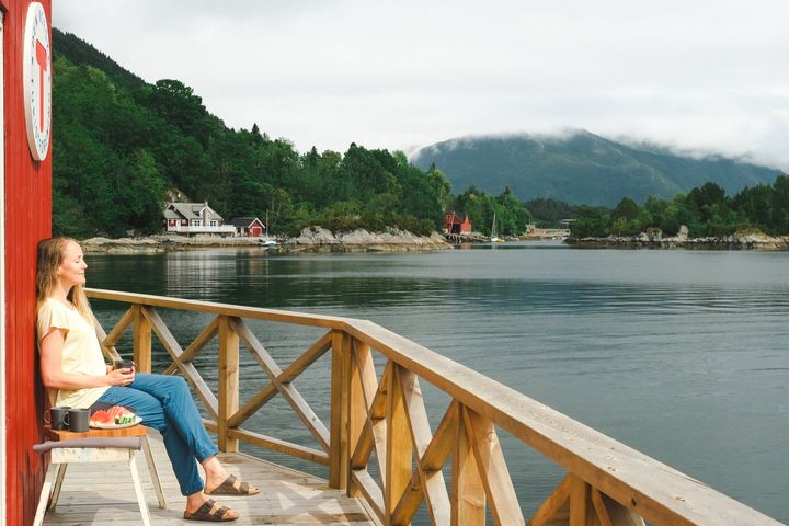 Kvinne sitter på brygge ved fjorden og nyter utsikten mot grønne skogkledde fjell.