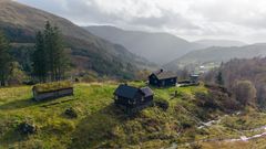 Luftfoto av små tømmerhus med torvtak på en grønn skråning i en norsk dal, omgitt av skog og fjell i disig lys.