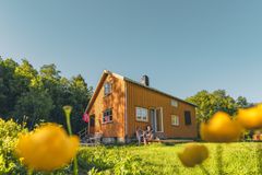 En gul trehytte med norsk flagg omgitt av grønn skog og gressletter med blomster. To personer sitter på trappen i kveldssol.