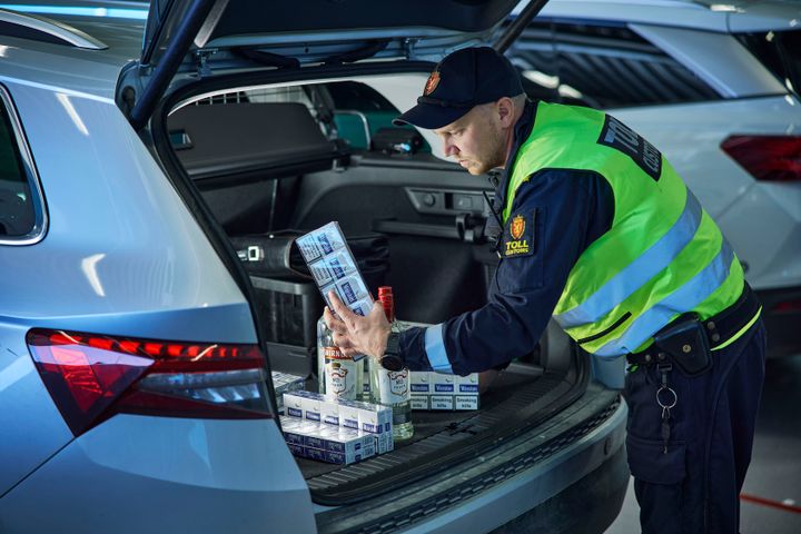 Toller i uniform med refleksvest står ved bagasjerommet på en bil, og undersøker flere røykpakker og brennevinsflasker.