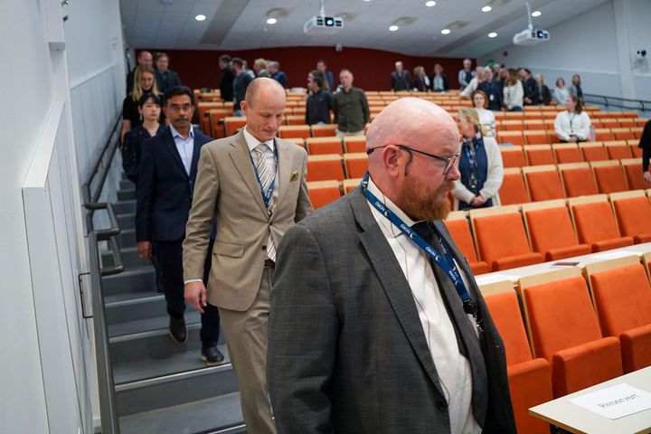 Flere voksne menn i dress som går ned en trapp i en forelesningssal.