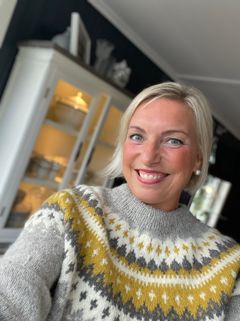 Selvportrett av kvinne med langt lyst hår og i en fargerik ullgenser, inne i sin egen stue.