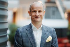 Bilde av voksen mann i lyseblå dress som smiler. Bildet er tatt inne, og i bakgrunn skimtes en rød sofa og en trappeoppgang.