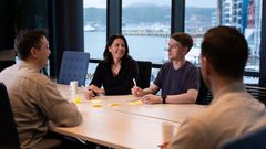 Fire voksne mennesker som snakker sammen rundt et møtebord, med utsikt mot ei havn i bakgrunnen.