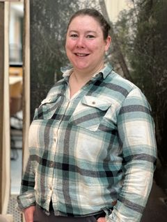 Portrett av voksen kvinne i rutete skjorte foran en vegg med bilde av trær.