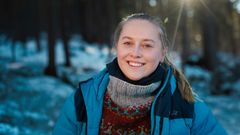 Ung kvinne ute i skogen en vinterdag med solstråler som trenger seg frem mellom trærne i bakgrunnen.