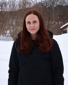 Portrettbilde av kvinne i svart jakke som står ute på en gårdsplass. Det er vinter og mye snø.