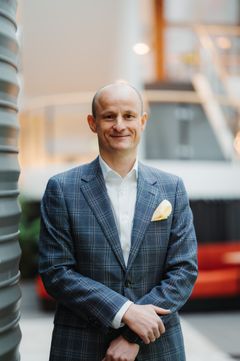 Voksen mann i dressjakke. Han står inne i en foaje.