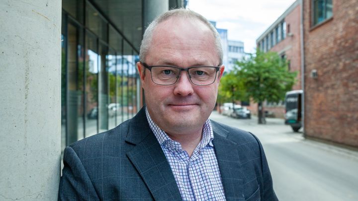 Voksen mann i dressjakke og skjorte. Han står ute i en gate ved en bygning. Det er oppholdsvær.