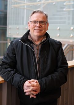 Voksen mann som står oppreist og ser i kamera. Han har sort jakke og foldede hender.