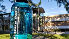 Blå vannflaske med samisk tekst på. I bakgrunnen et universitetsbygg og blå himmel.