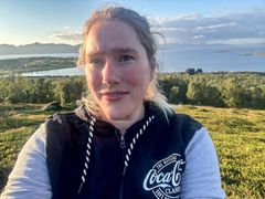 Selvportrett av ung kvinne. Hun står ute på en grønn slette. I bakgrunnen ser vi fjord og fjell. Det er pent vær.