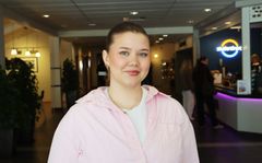 Ung kvinnelig student i rosa genser. Hun står inne i en foaje.