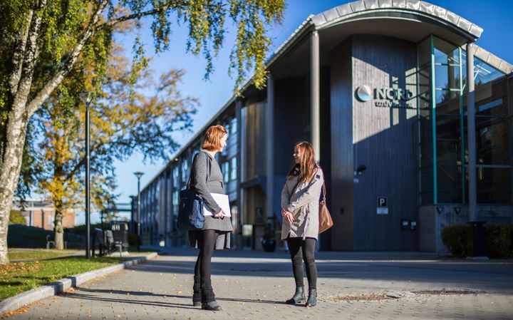 To voksne mennesker foran universitetsbygg. Det er sommer og blå himmel.