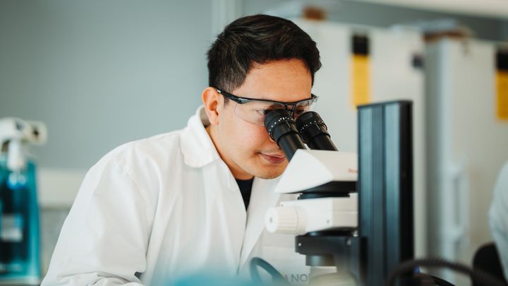 En person i laboratoriefrakk ser gjennom et mikroskop.