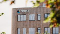Utsnitt av bygning med logo og vinduer. I forgrunnen grønne blader og greiner.