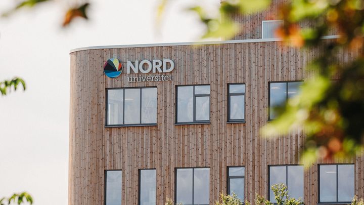 Utsnitt av bygning med logo og vinduer. I forgrunnen grønne blader og greiner.