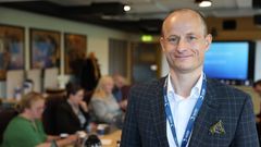 Voksen mann i dress som står inne i et møterom med andre møtedeltakere i bakgrunnen rundt et bord