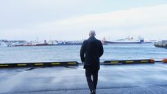 Vi ser ryggen på en voksen mann som går på en kai. I bakgrunnen ser vi havet og flere skip.