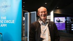 En voksen mann ser i kamera og smiler. Han er inne i et konferanseområde, og står ved siden av et beachflagg for konferansen.