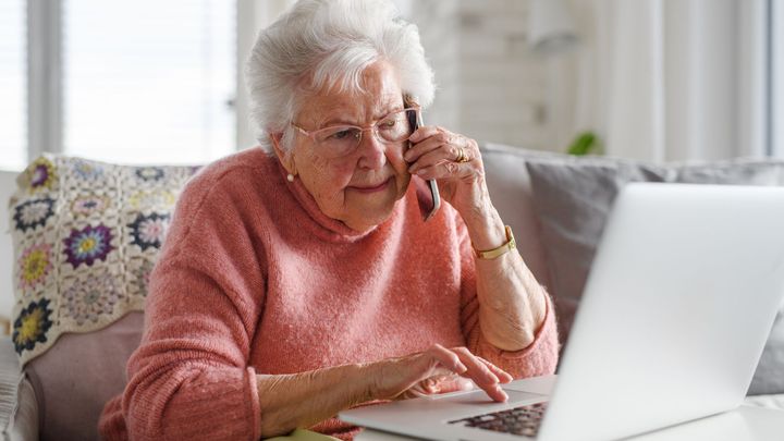 Eldre kvinne sitter i sofaen i mobiltelefonen samtidig som hun taster på en bærbar datamaskin