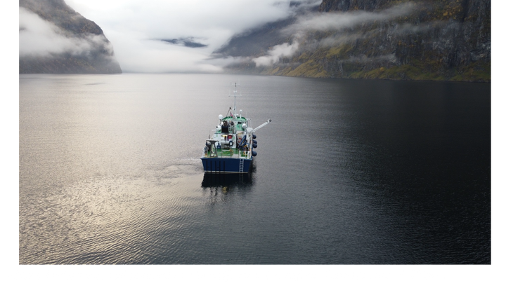 Dronefoto av skipet «Hans Brattstrøm» som seiler inn i Nærøyfjorden mellom høye, tåkelagte fjell.