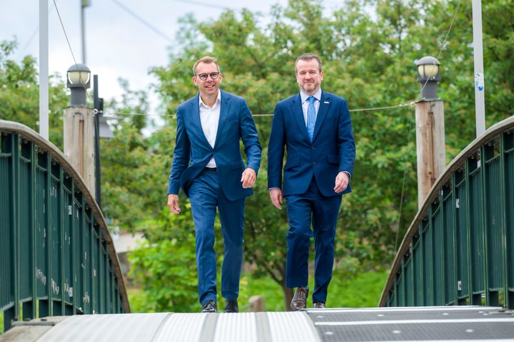 To menn som går over en bro. Trær, himmel, lyktestolper. Foto.