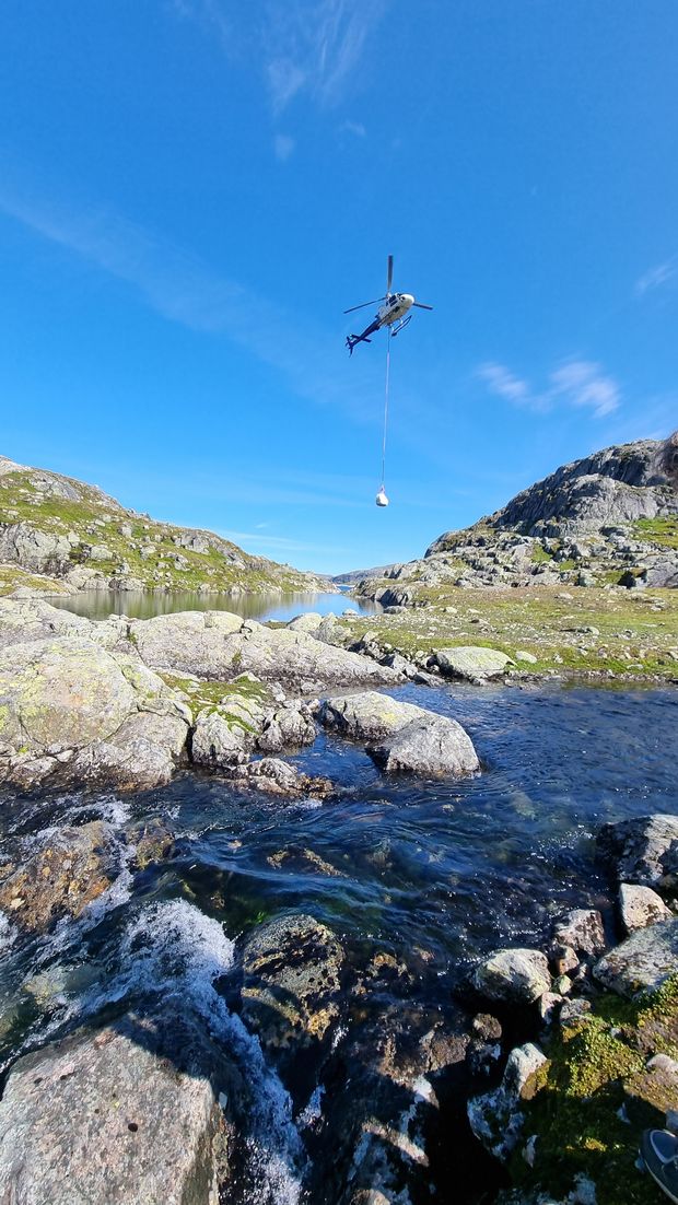 Helikopter, himmel, fjellterreng, bekk, grus. Foto.