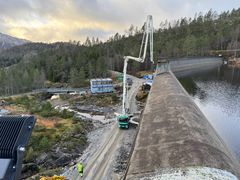 Dam, anleggsmaskiner, anleggsbrakker, vann, skog, himmel. Foto.