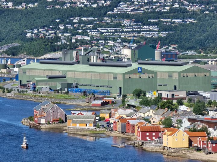 Bilde av Alcoas aluminiumsverk og bebyggelse rundt. Verket ligger ved kysten. I forgrunnen ligger gammel trebebyggelse langs kystlinjen. I bakgrunnen er det skogkledd fjell og nyere bebyggelse.