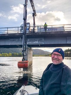 Mann i båt, lastebil på bro som legger ut grus i elva. Foto.