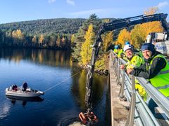 Menn i arbeidstøy og lastebil på bro, to menn i båt under broa, lastebil legger ut grus i elva. Foto.