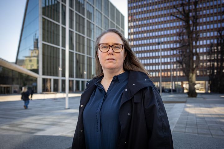 Kvinne med brunt hår og brune briller, mørkeblå bluse og svart jakke foran bygninger i regjeringskvartalet.