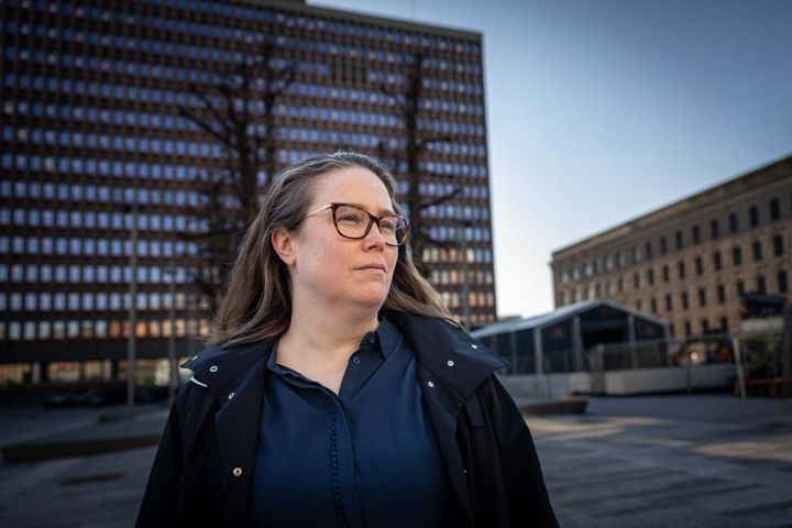 Kvinne med brunt hår og brune briller, mørkeblå bluse og svart jakke foran bygninger i regjeringskvartalet.