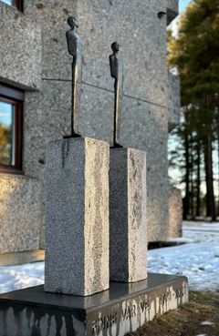 To bronseskulpturer står på hver sin granittsokkel, som igjen er montert sammen i en betongsokkel med tittelen 'Finne vår vei' støpt inn i betongen.