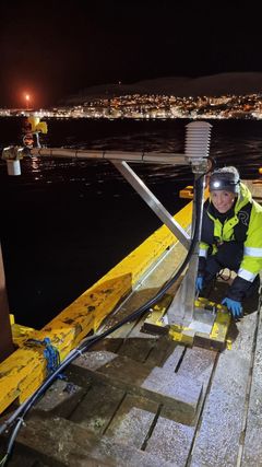 Bilde av en person som reparerer en vannstandmåler ved sjøen i nattens mørke, med lys fra byen Hammerfest i bakgrunnen.