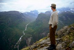 En person står på en fjelltopp og ser ut over en dal, fjell og ei elv i det fjerne.