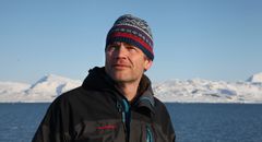 Halfdan Pascal Kierulf er godt kledt i jakke og lue. Bak ham er hav og snødekte fjell i Ny-Ålesund.