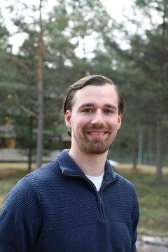 Carl William Lund smiler. Han har på seg ei blå skjorte og det er skog i bakgrunnen.