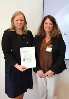 To kvinner står ved siden av hverandre. Den ene holder viser fram en rapport.