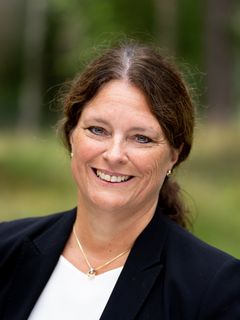 Ei smilende kvinne med brunt hår og en svart jakke står utendørs.