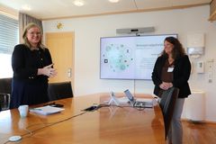 To kvinner står ved et møterom med en presentasjon på skjermen i bakgrunnen.