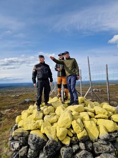 Tre menn står på toppen av en gulmalt riksrøys ved en riksgrensa, hvorav en peker utover landskapet.