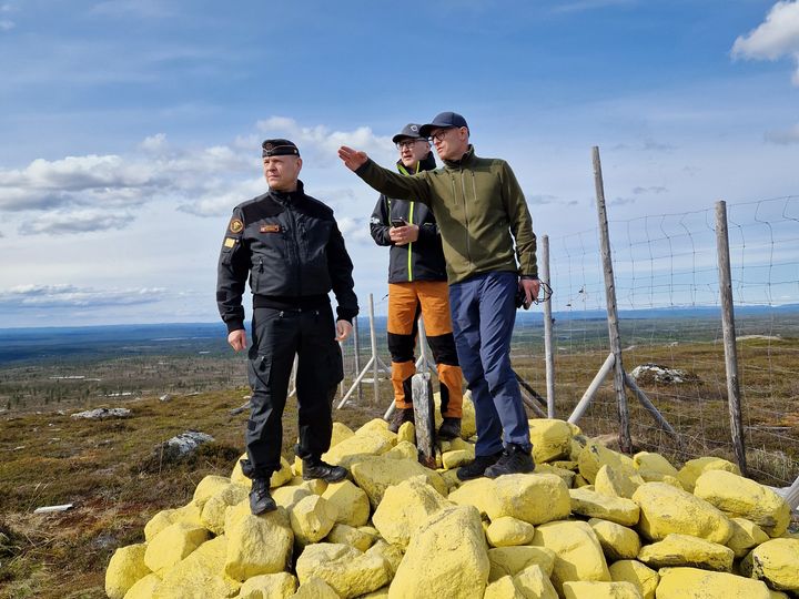 Tre menn står på toppen av en gulmalt riksrøys ved en riksgrensa, hvorav en peker utover landskapet.