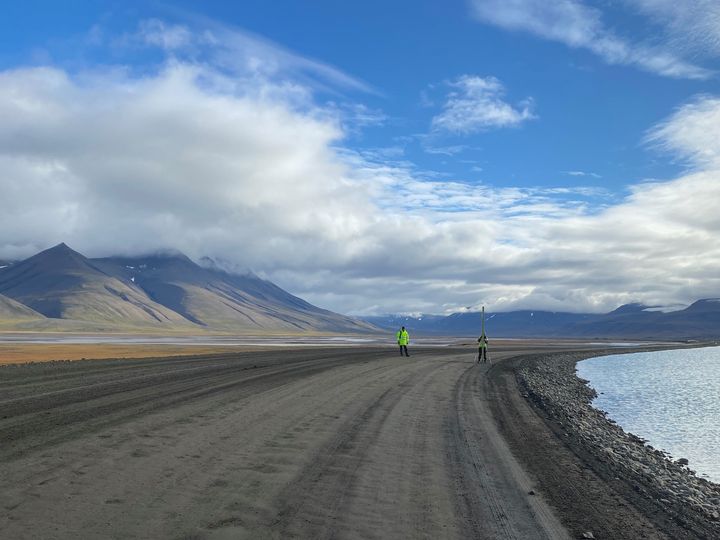 To personer i gule jakker utfører feltarbeid langs en grusvei ved siden av hav, med fjellandskap i bakgrunnen under en skyet himmel.