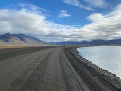 To personer i gule jakker utfører feltarbeid langs en grusvei ved siden av hav, med fjellandskap i bakgrunnen under en skyet himmel.