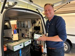 En mann står ved siden av en bil fylt med teknisk utstyr, og holder GNSS i hendene.