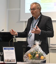 En person står og gir en presentasjon ved et bord. På bordet er det en blomstergave og et innrammet dokument.