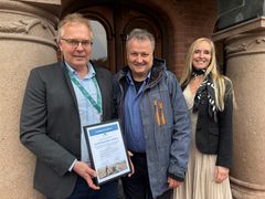Tre personer står utenfor en bygning, den ene holder et innrammet