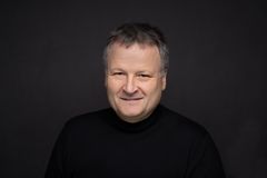 Mann med mørk genser og kort hår smiler i portrettfoto mot mørk bakgrunn.